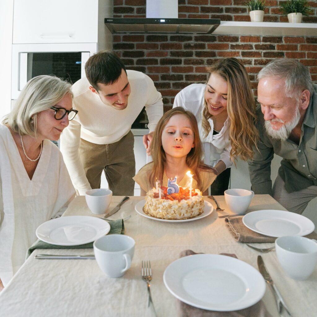A young girl blows out candles on her birthday cake surrounded by family members indoors.